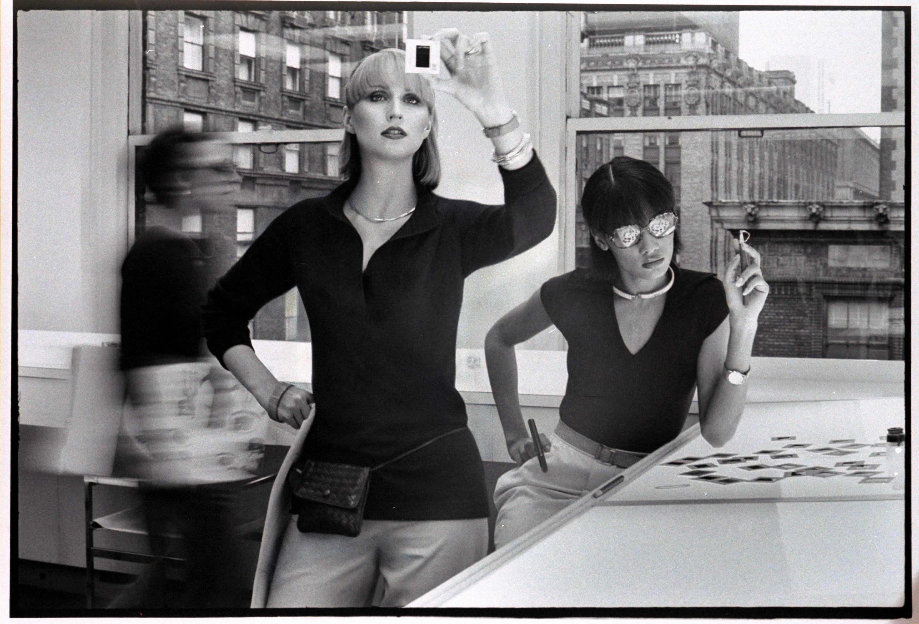 Vogue, February 1976 - Two models in an office looking at negatives, and wearing Kimberly Sport ensembles: Model (Chris Royer), left, is wearing a dark pullover, slim white pants with small purse around her waist; Right, model is wearing reading glasses, dark fitted pullover with cap sleeves and belted off-white skirt. (Duane Michals/Conde Nast via Getty Images)