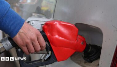 A close-up image of a silver car being filled up at a fuel pump, the pump is red and black. The person holding the pump is wearing a blue jumper.