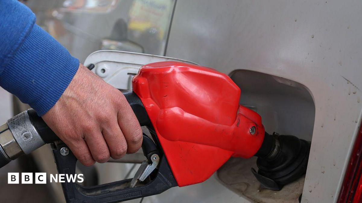 A close-up image of a silver car being filled up at a fuel pump, the pump is red and black. The person holding the pump is wearing a blue jumper.