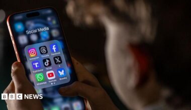A child, who is out of focus, looks at a phone held in both hands. On the screen is a folder labelled 'social media' with app icons for Instagram, Threads, Facebook, TruthSocial, TikTok, X, WhatsApp, YouTube and Bluesky.