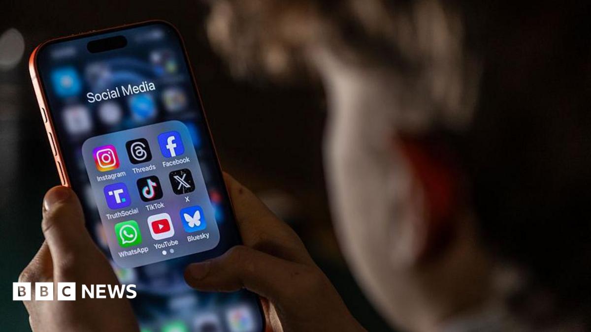 A child, who is out of focus, looks at a phone held in both hands. On the screen is a folder labelled 'social media' with app icons for Instagram, Threads, Facebook, TruthSocial, TikTok, X, WhatsApp, YouTube and Bluesky.