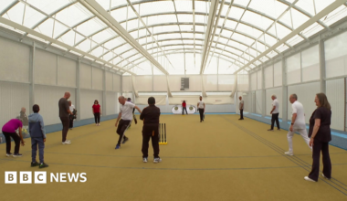 A large sports hall with white walls, an arched roof and yellow floor with people standing around and cricket stumps in the foreground