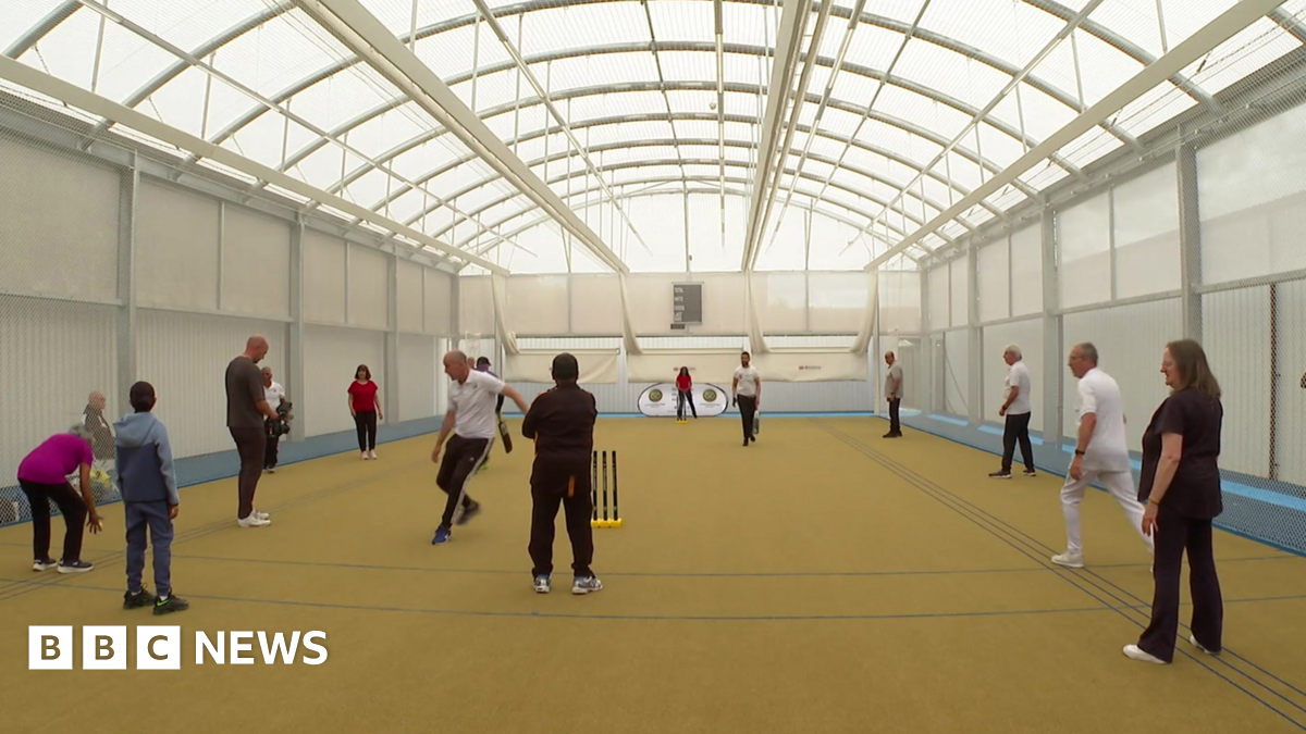 A large sports hall with white walls, an arched roof and yellow floor with people standing around and cricket stumps in the foreground