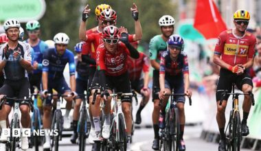 Geraint Thomas crosses the finish line cheered by the riders in his farewell and last race as a professional cyclist during the 21st Tour of Britain 2025. The riders are all wearing cycling colours and helmets.