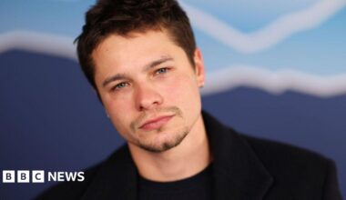 Toby is looking into the camera, he has a black t-shirt on with a black jacket over the top. He has short brown hair and blue eyes.