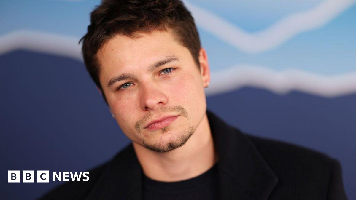 Toby is looking into the camera, he has a black t-shirt on with a black jacket over the top. He has short brown hair and blue eyes.
