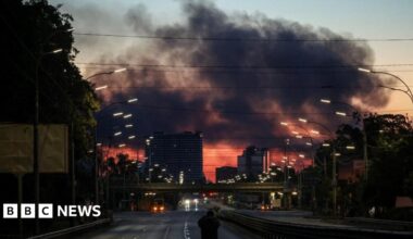 A man stands on an empty highway to photograph dark clouds of smoke rising through the early morning sky over a city.