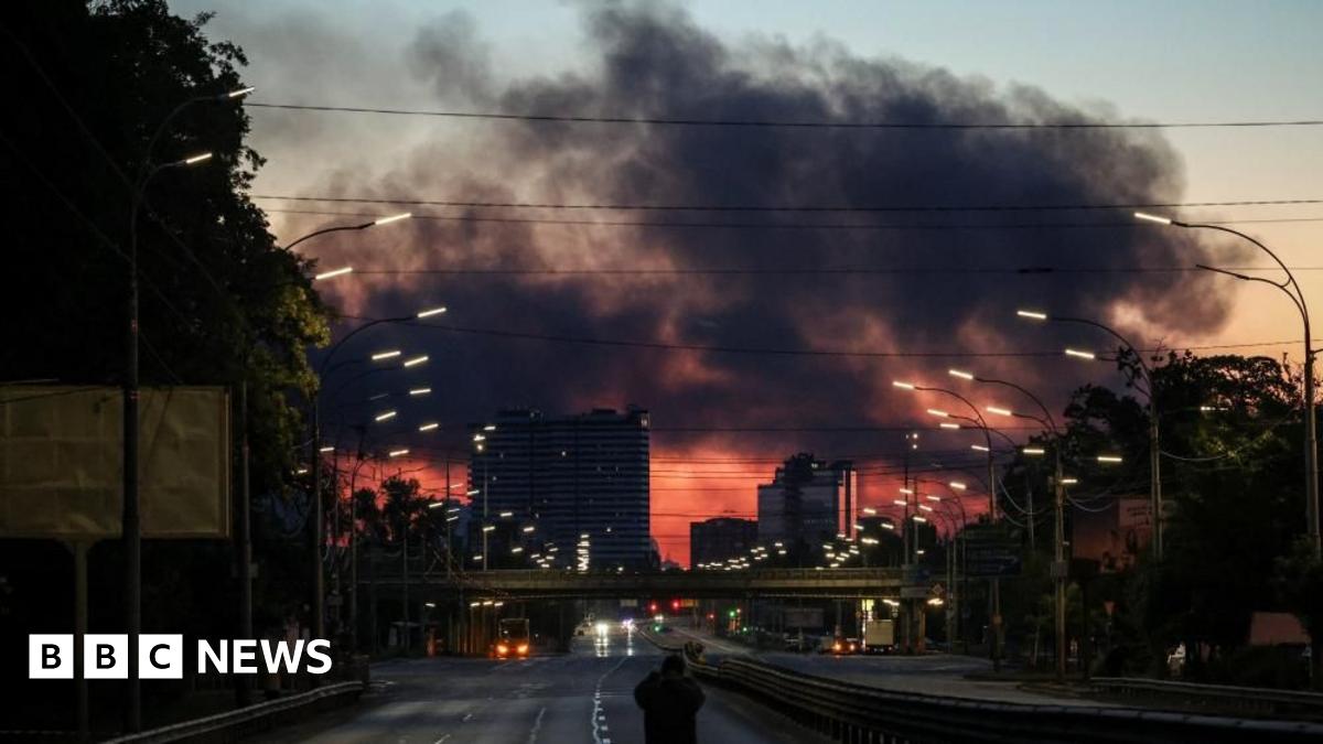 A man stands on an empty highway to photograph dark clouds of smoke rising through the early morning sky over a city.