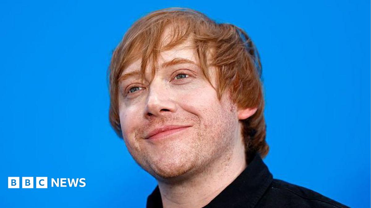 Rupert Grint is stood against a blue background and smiling. He has auburn hair and is wearing a black shirt.