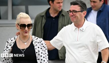 Marie-France van Heel pictured next to her husband, Andy Burnham. She is wearing a black top with a white shirt with black polka dots over the top. She has sunglasses and necklaces. Andy Burnham has his arm around her and is wearing a white polo shirt.