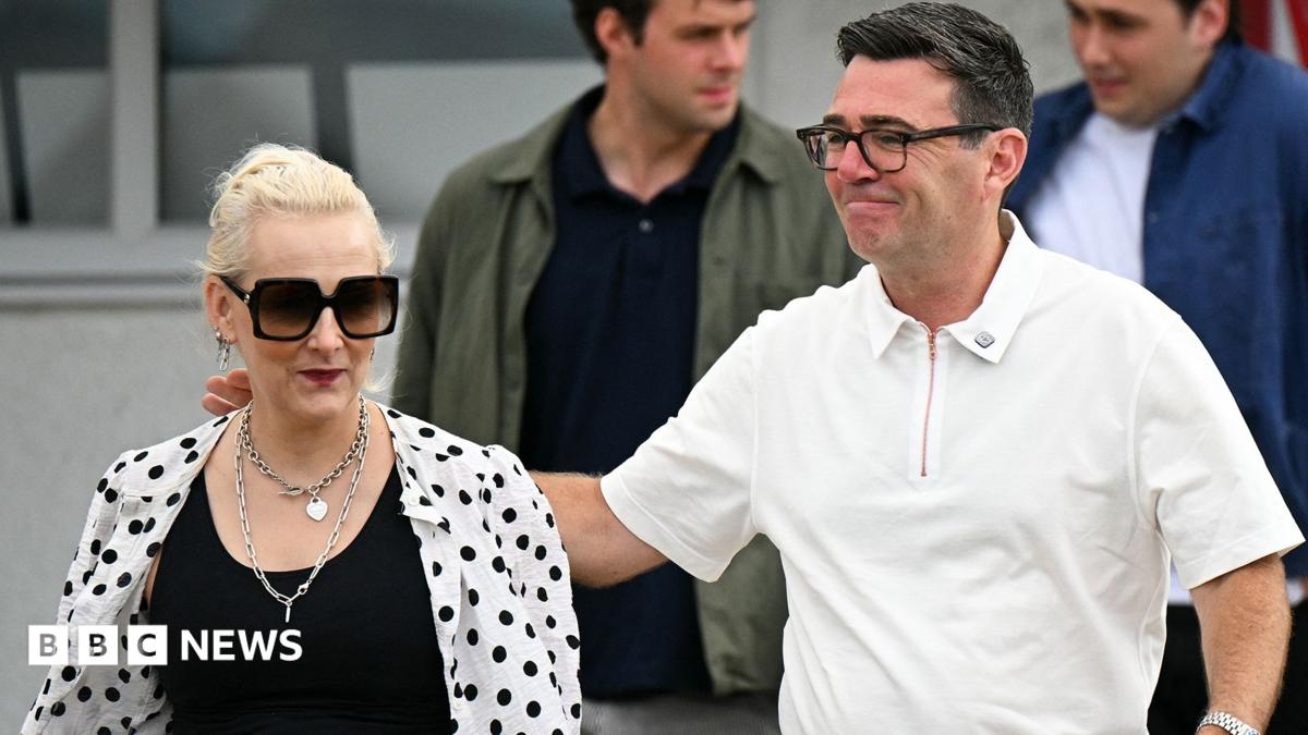 Marie-France van Heel pictured next to her husband, Andy Burnham. She is wearing a black top with a white shirt with black polka dots over the top. She has sunglasses and necklaces. Andy Burnham has his arm around her and is wearing a white polo shirt.