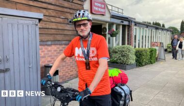 A man on a bicycle, wearing a helmet, gloves, a lanyard and a red T-shirt reading MND Association.