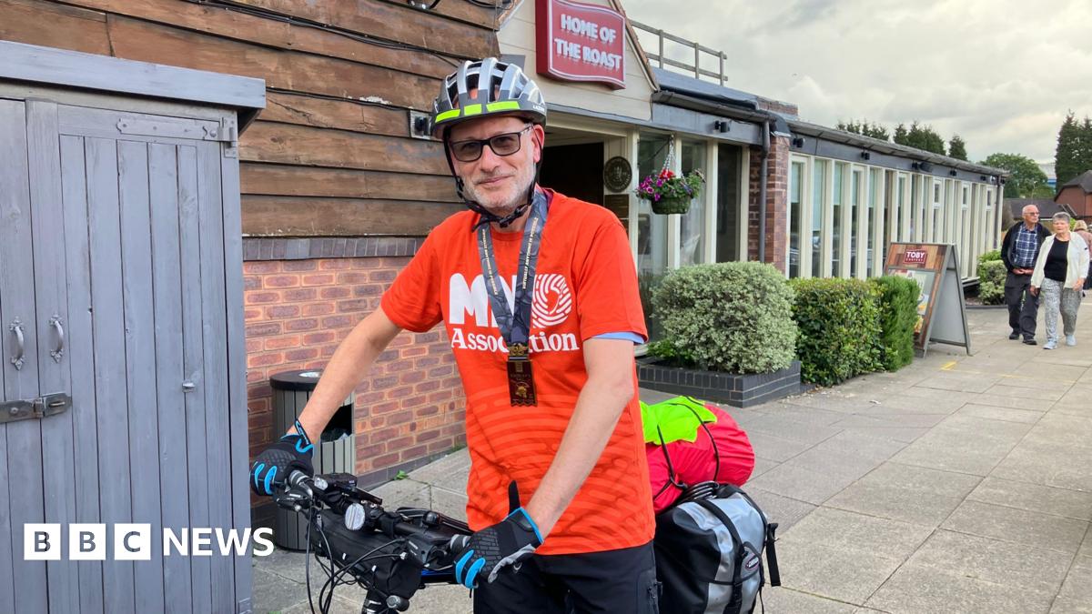 A man on a bicycle, wearing a helmet, gloves, a lanyard and a red T-shirt reading MND Association.
