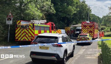 A fire engine is parked on the left side of a road with a fire and rescue car on the other side. There is a truck with Command Unit written on the back in the background and police tape is cordoning off the scene.