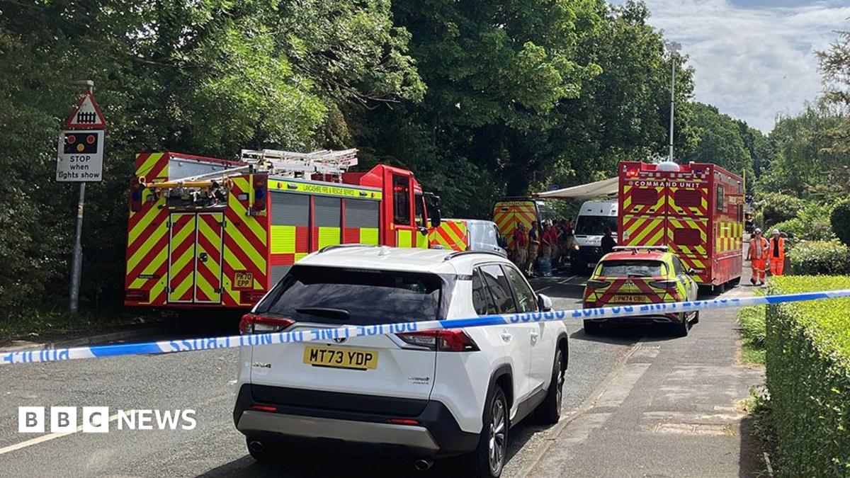 A fire engine is parked on the left side of a road with a fire and rescue car on the other side. There is a truck with Command Unit written on the back in the background and police tape is cordoning off the scene.