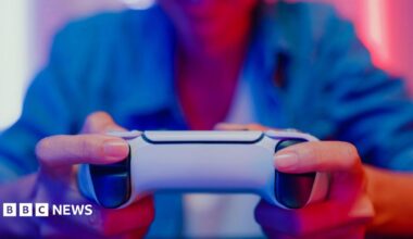 Close up of a woman's hands gripping a gaming controller, with colourful lighting reflecting on her from a monitor screen in front.