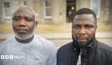 Two men shown side-by-side in a split portrait, both facing the camera outdoors in front of a blurred building. The man on the left has a short haircut with a grey beard and is wearing a light-coloured patterned shirt, while the man on the right has short hair, a trimmed beard, and is wearing a dark quilted jacket over a shirt.