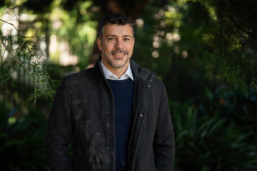 Cliff wearing a warm jacket over a collared shirt and jumper stands in front of greenery, smiling.