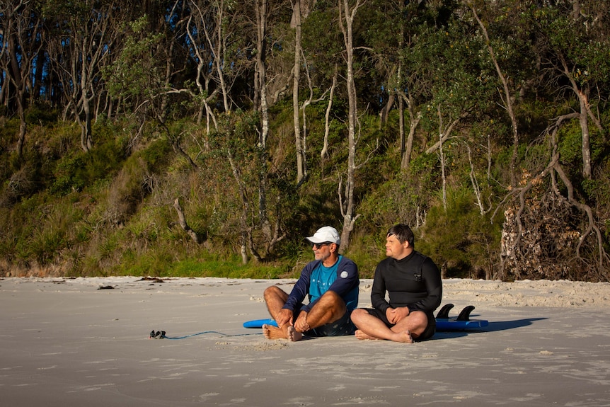 A young man and an older man sit on a beach in front of a surfboard in early light