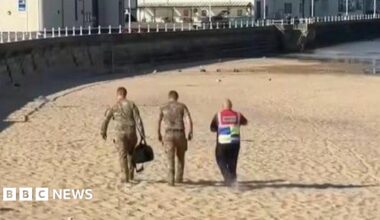 Three people are walking along a sandy beach with seafront buildings in the background. Two are wearing camouflage with one carrying a large black bag, while the third is wearing a high-vis vest.