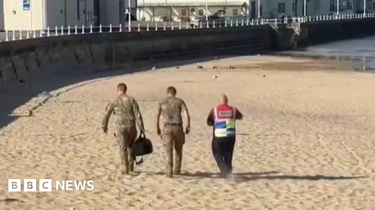Three people are walking along a sandy beach with seafront buildings in the background. Two are wearing camouflage with one carrying a large black bag, while the third is wearing a high-vis vest.