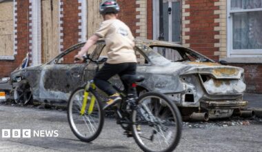 A burnt out car is on a street. Boarded up terraced houses are behind it. There is a kid on a bike in front of the burnt-out car.