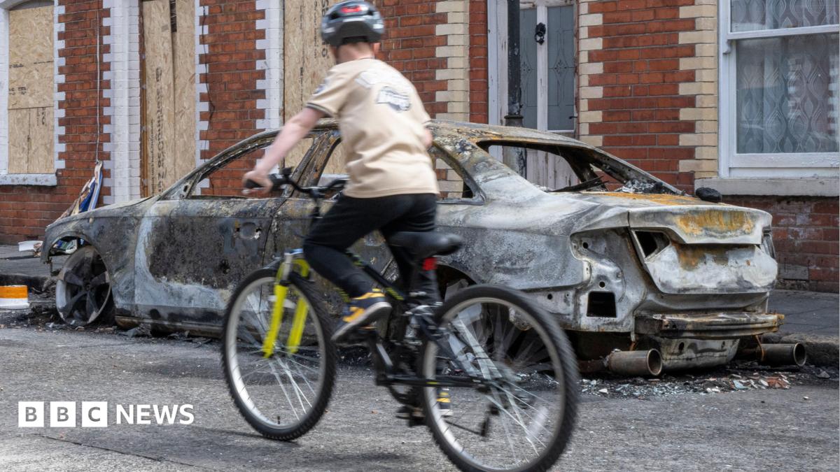 A burnt out car is on a street. Boarded up terraced houses are behind it. There is a kid on a bike in front of the burnt-out car.