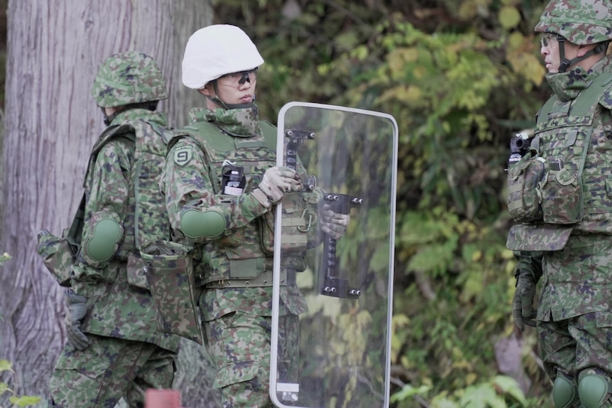 A man in a white helmet and military fatigues with a large transparent shield standing near some trees.