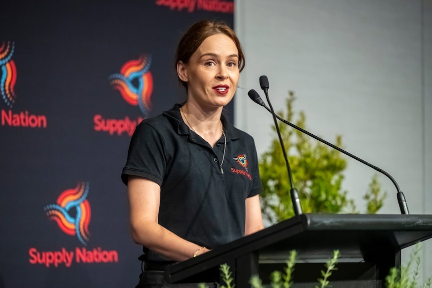 Kate is wearing a black supply nation shirt standing in front of a lecturn