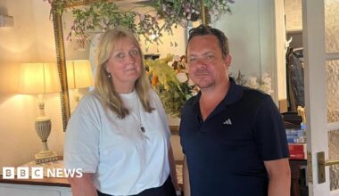 A women wearing a white t-shirt stood with a man wearing a blue polo shirt. They are both looking at the camera.