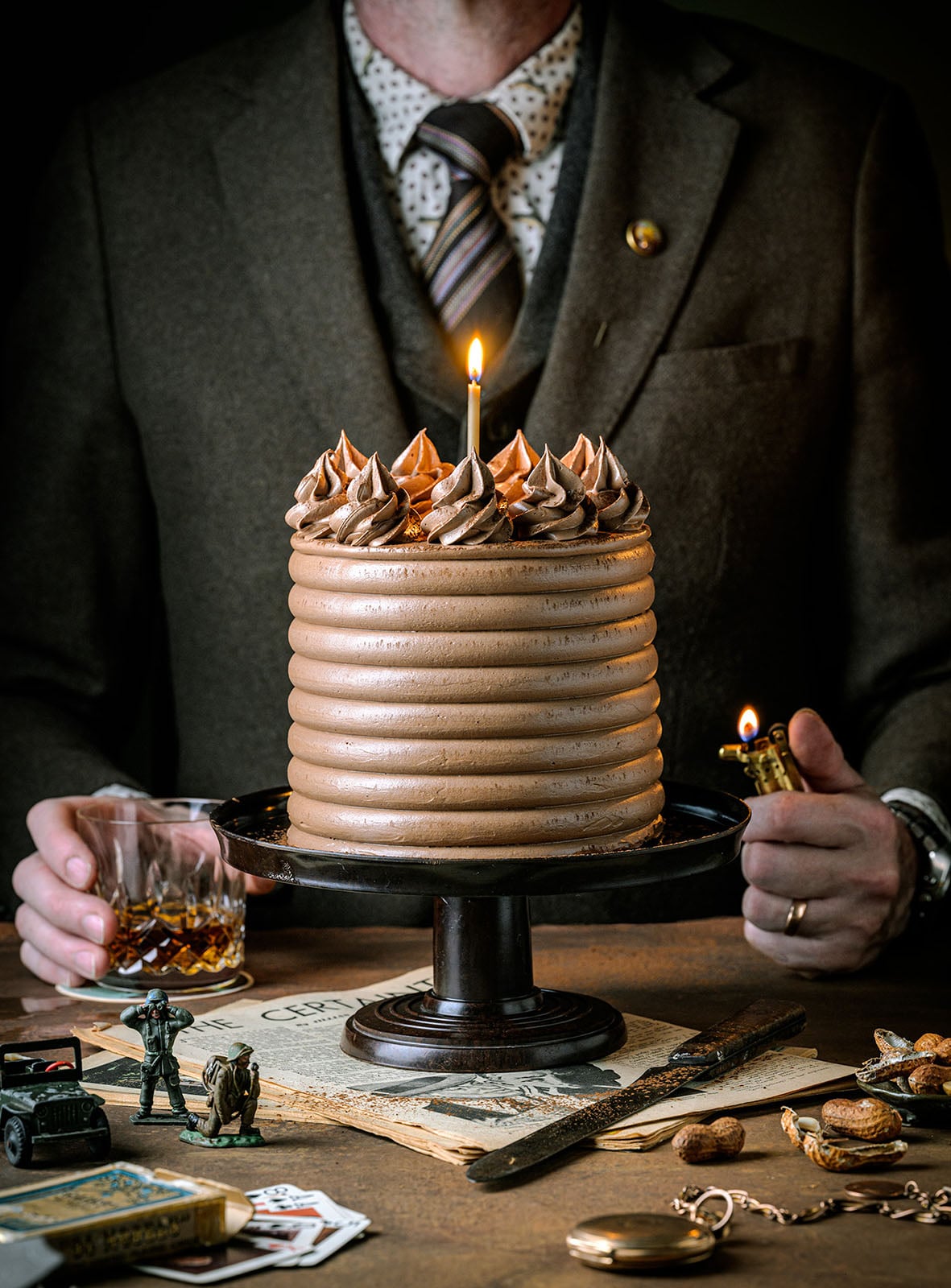 A person in a brown suit holds a glass of whiskey and a lighter behind a tall, frosted chocolate cake with a lit candle on a stand; vintage items and toys are scattered on the table.