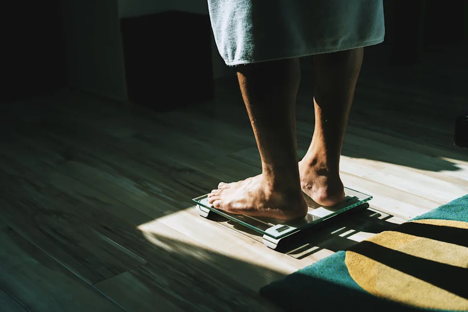 Person standing on a bathroom scale with soft lighting. The scene suggests a focus on personal health or weight monitoring