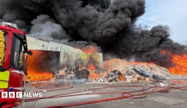 The front of a fire engine can be seen on the left of the image, with a large pile of cardboard on fire and large plumes of smoke billowing into the sky. A single-storey building can also be seen ablaze.