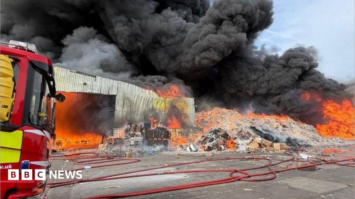 The front of a fire engine can be seen on the left of the image, with a large pile of cardboard on fire and large plumes of smoke billowing into the sky. A single-storey building can also be seen ablaze.