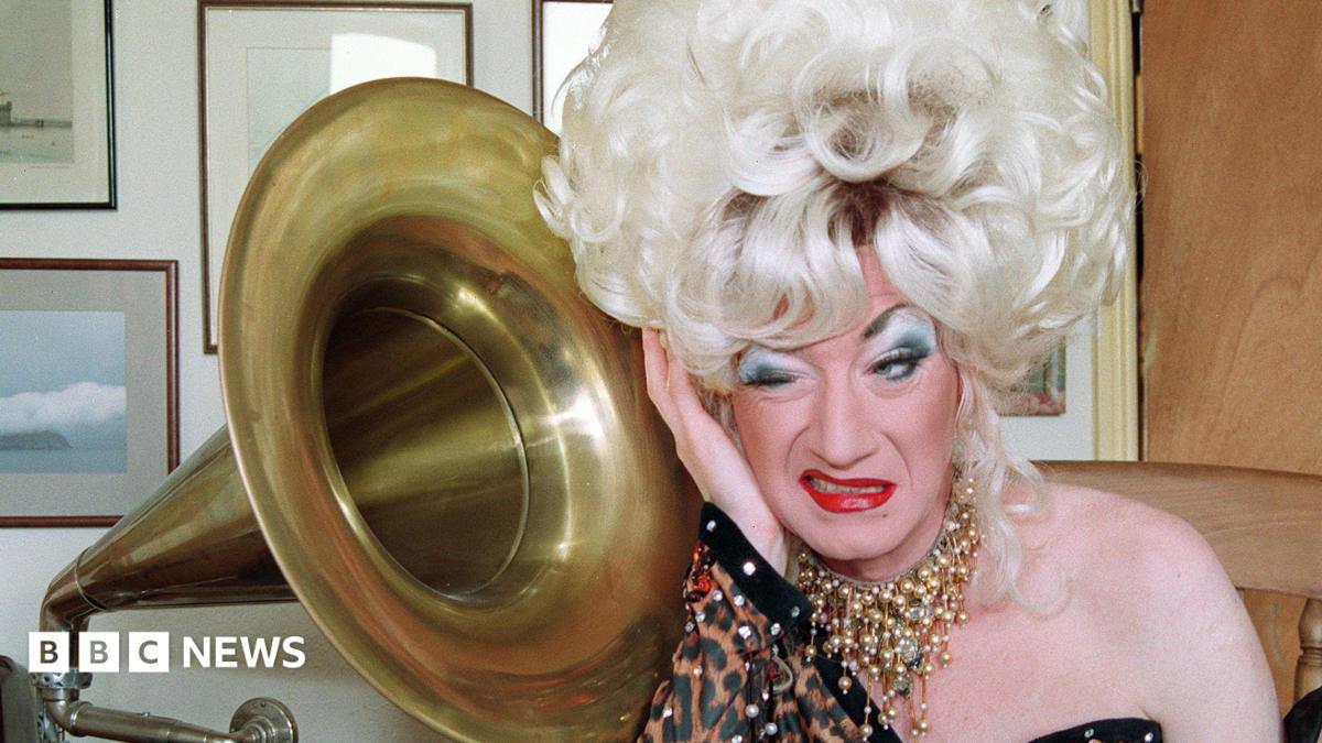 Paul O'Grady as his alter ego Lily Savage in a blonde wig next to a grammarphone