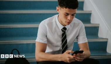 A teenage boy in school, wearing a shirt and tie, sat on some stairs, using his phone