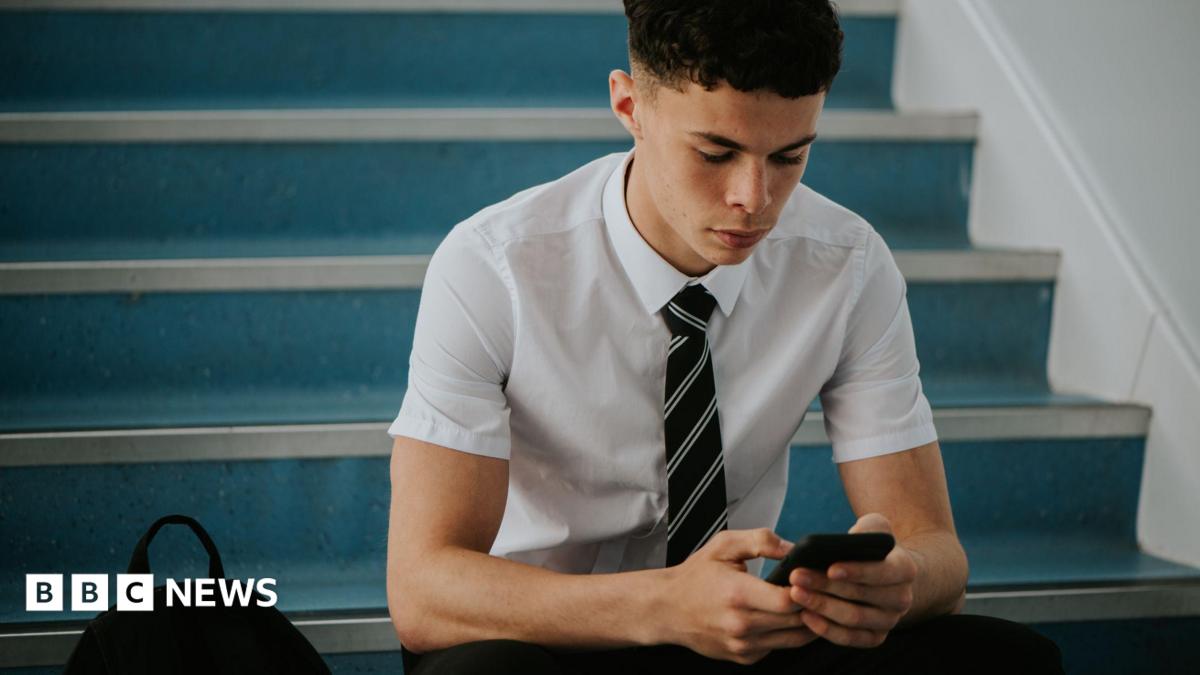 A teenage boy in school, wearing a shirt and tie, sat on some stairs, using his phone
