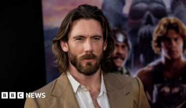 James Wilkinson. He has long dark hair and a beard. He is wearing a tan suit jacket and an open white shirt. A blurred movie poster is behind him.
