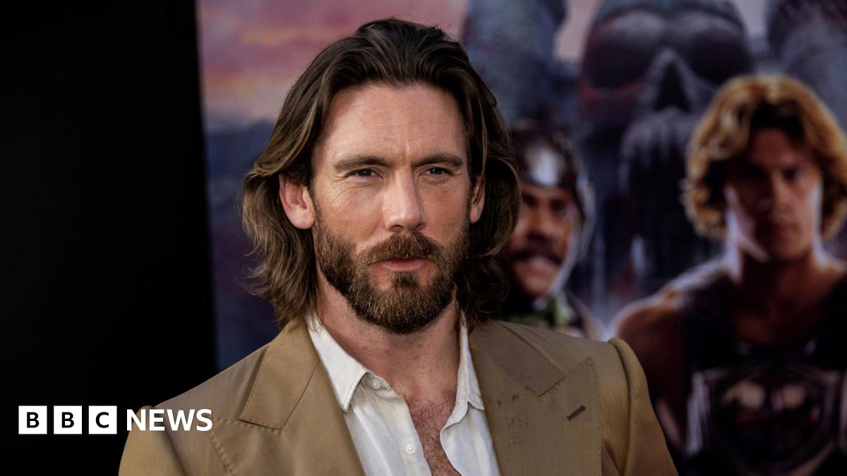 James Wilkinson. He has long dark hair and a beard. He is wearing a tan suit jacket and an open white shirt. A blurred movie poster is behind him.