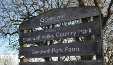 A mainly dark sign that includes the words Welcome to Sandwell Valley Country Park. Trees and grass are in the background.