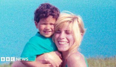 A young child with curly hair is being held closely by an adult with light-coloured hair; both are positioned outdoors against a bright blue sky, with grassy landscape visible in the background.