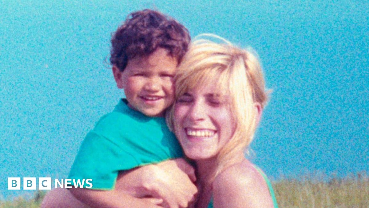 A young child with curly hair is being held closely by an adult with light-coloured hair; both are positioned outdoors against a bright blue sky, with grassy landscape visible in the background.