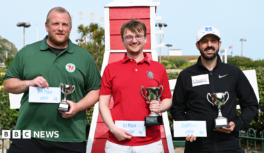 The image shows three men standing with trophies and certificates showing their podium placement. The winner, in the middle, is wearing a red shirt, the man in 2nd place on the right is wearing a black shirt and the last finalist on the left is wearing a green shirt. Behind them, bushes can be seen.