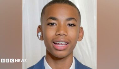 Head shot of Jamal Coombes who is wearing a white shirt beneath a blue jacket. He has a white air pod in one ear