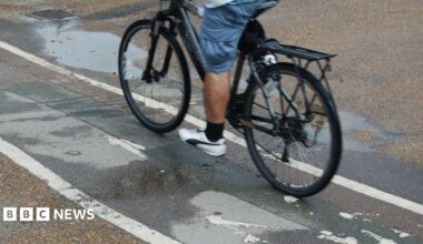 A man cycling on a cycle path. Only the lower half of his body is visible. He is wearing blue shorts, black socks and white trainers.