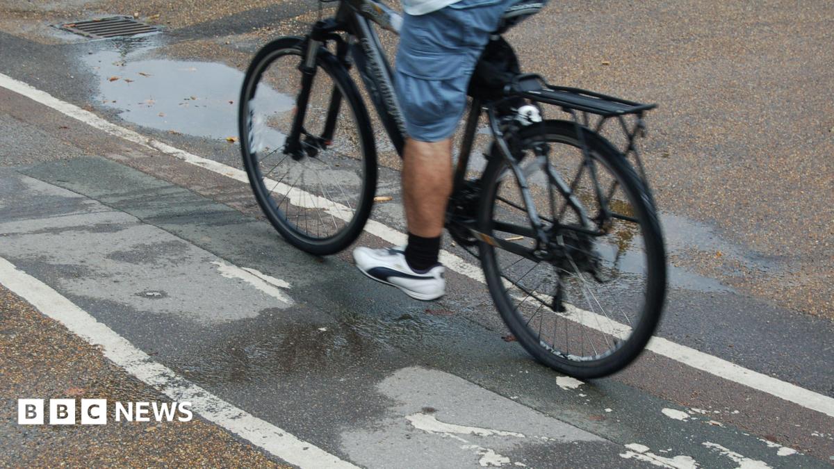 A man cycling on a cycle path. Only the lower half of his body is visible. He is wearing blue shorts, black socks and white trainers.