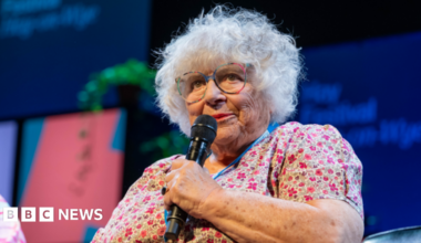 Close up of Margoyles talking on stage at Hay Festival. She wears a floral dress, colourful glasses and has short curly grey hair. She holds a black microphone and looks out at audience.