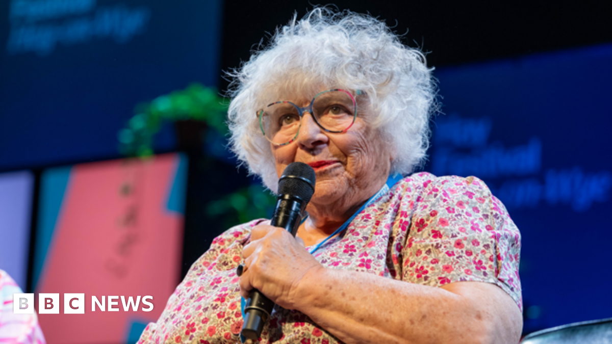 Close up of Margoyles talking on stage at Hay Festival. She wears a floral dress, colourful glasses and has short curly grey hair. She holds a black microphone and looks out at audience.