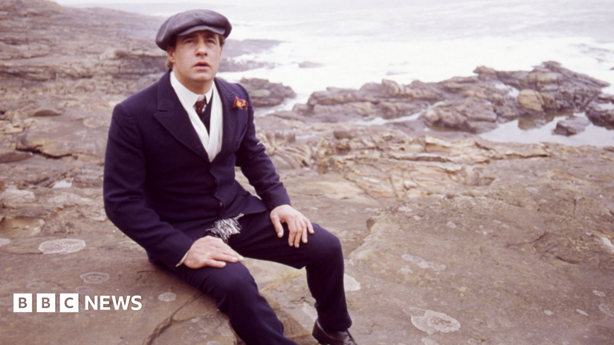 David appearing in Centre Play: God Bless Thee Jacky Maddison - Play by Tom Hadaway. Picture shows David Daker as Jacky Maddison. He is sat on rocks on a beach with a hat on.