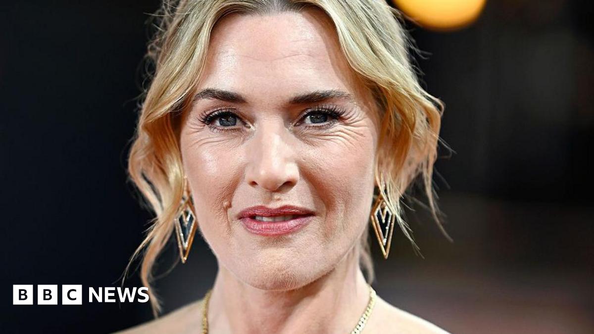 A head and shoulders image of actress Kate Winslet. She has short blonde hair and is wearing a gold necklace.